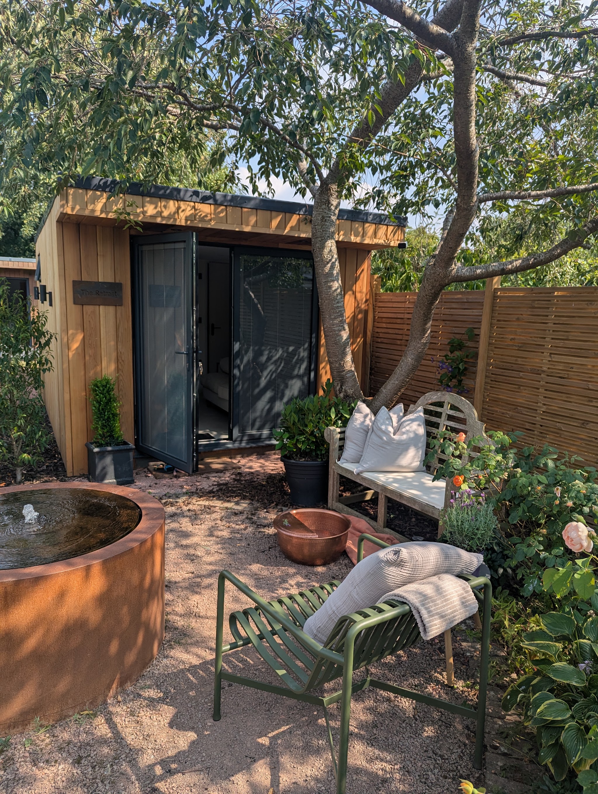 The timber-clad garden studio with its doors open, a corten water feature, a copper foot bowl and garden seating